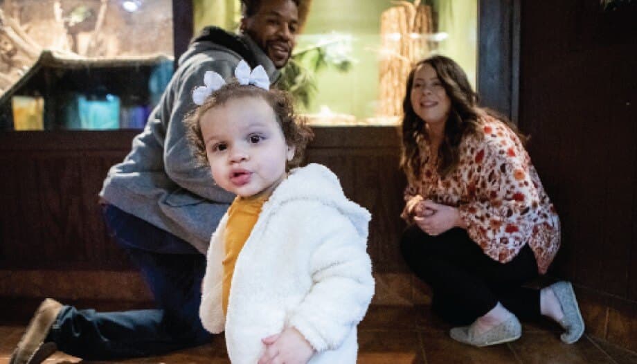 A toddler girl looking forward as her parents gaze at her with pride