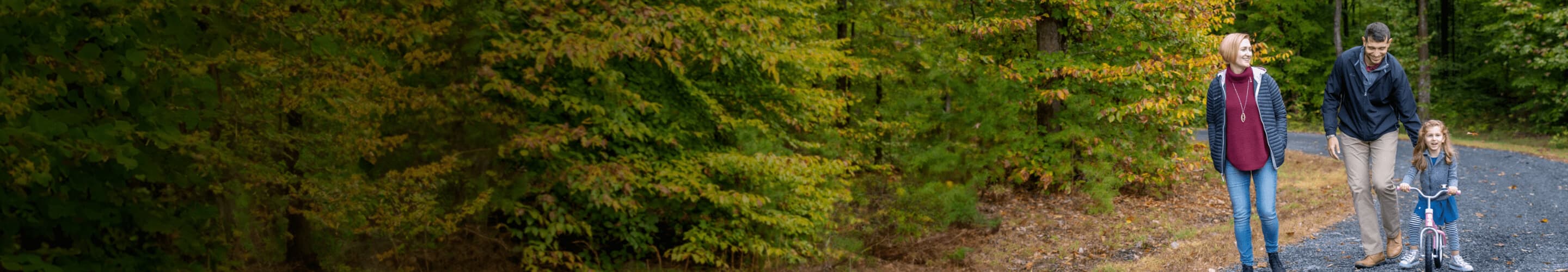 Parents accompanying their young child, who is bicycling on a leafy path