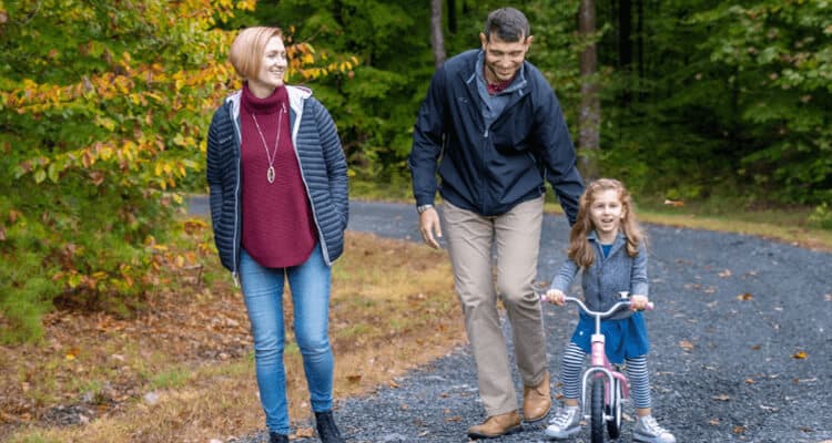 Parents accompanying their young child, who is bicycling on a leafy path
