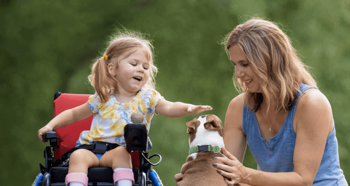 Mom with toddler girl in wheelchair, who is eagerly petting a dog