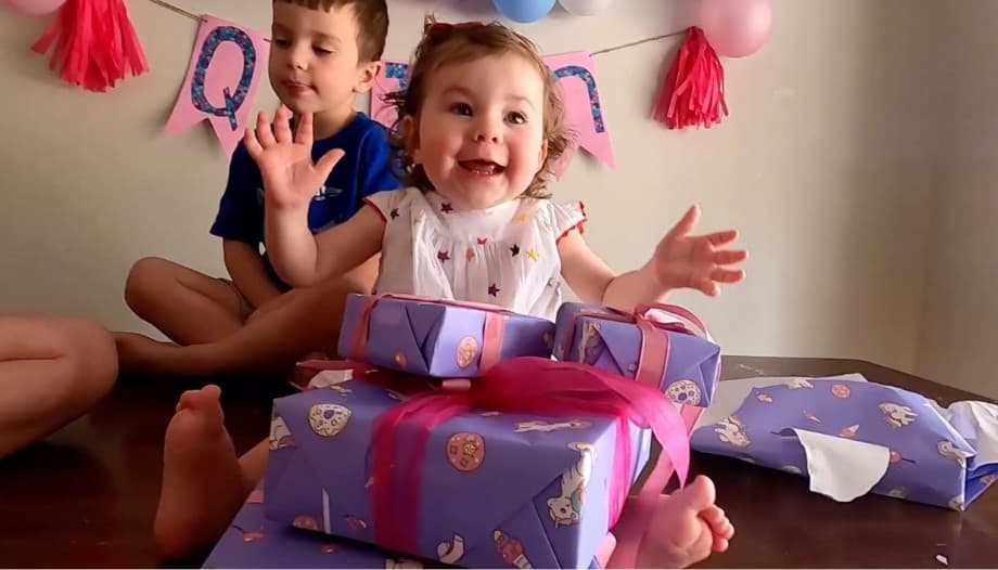 Video: an excited toddler girl sits in front of a pile of presents as her brother looks on